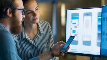 Due persone analizzano interfacce app su schermo e telefono.