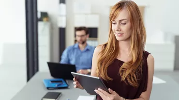 Persona in ufficio con tablet, sfondo sfocato con collega al computer.