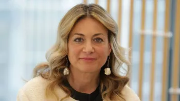 Persona sorridente con capelli ondulati e sfondo di finestre e legno.
