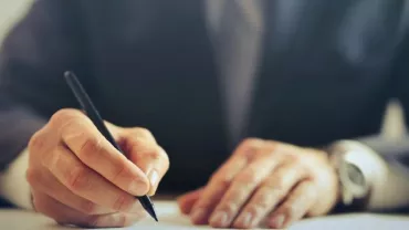 Persona con abito formale scrive su un foglio con una penna.