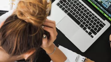 Persona stressata lavora al computer portatile con appunti e occhiali da vista sul tavolo.
