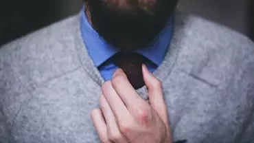 Persona sistema una cravatta rossa su camicia blu e maglione grigio.