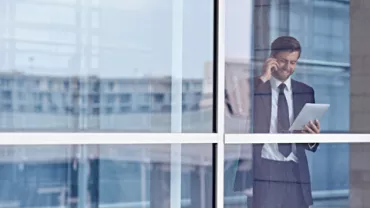 Persona che parla al telefono e guarda un tablet, vista attraverso una finestra.