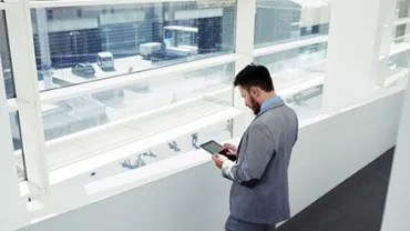 Persona in abito grigio guarda un tablet vicino alla finestra.