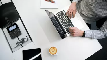 Persona lavorando al computer con caffè, telefono e tablet su un tavolo bianco.