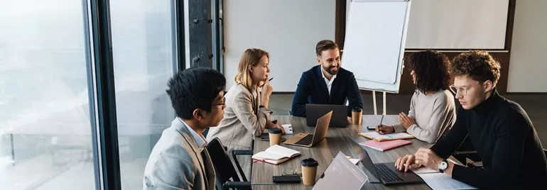 Gruppo in riunione attorno a un tavolo con laptop e appunti.