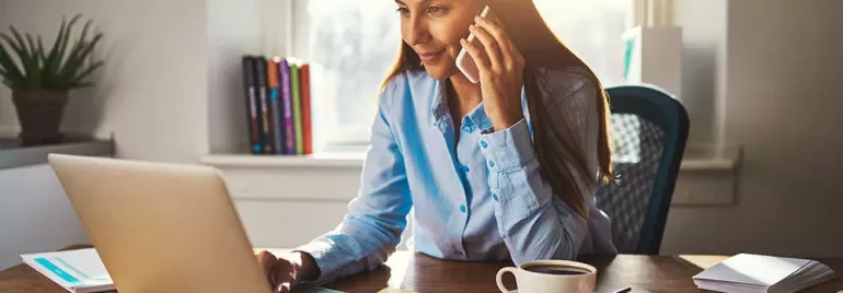 Persona al telefono mentre lavora al computer portatile in un ufficio illuminato dal sole.