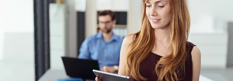 Persona in ufficio con tablet, sfondo sfocato con collega al computer.