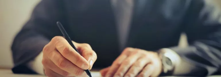 Persona con abito formale scrive su un foglio con una penna.