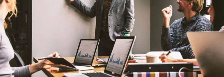 Riunione in ufficio con persone che discutono e laptop aperti sul tavolo.