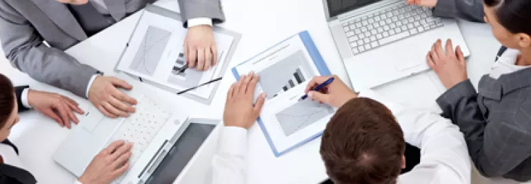 Persone in riunione, laptop e grafici sul tavolo.