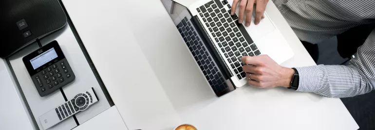 Persona lavorando al computer con caffè, telefono e tablet su un tavolo bianco.