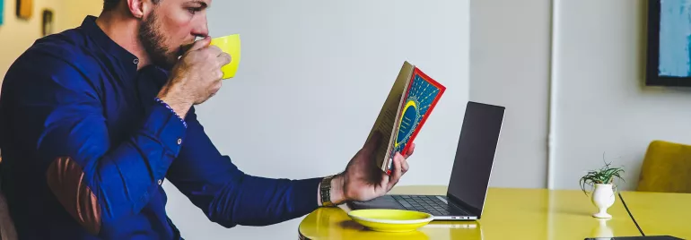 Persona seduta con tazza, libro aperto e laptop su tavolo giallo