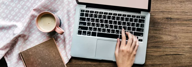 Persona usa un laptop con una tazza di caffè e un libro su tavolo di legno.